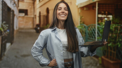 Woman holding laptop with one hand and hand on hip in a narrow urban street alleyway smiling at...