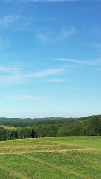 Wide view of green rolling fields and forested hills in the Morvan countryside on a clear summer day. Expansive blue sky with light clouds above rural landscape.