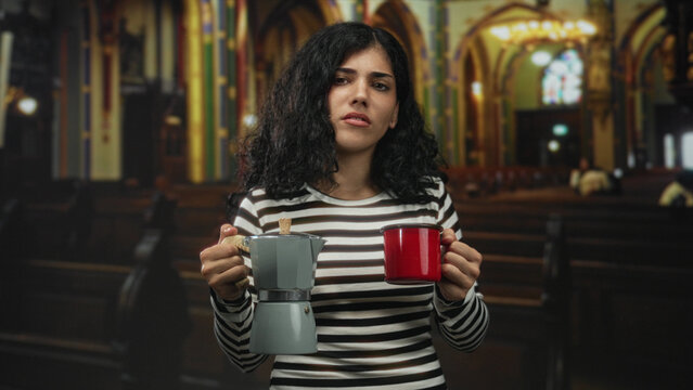 Young arab brunette woman holds stovetop coffee pot and red enamel mug in church building; ritual routine confusion.