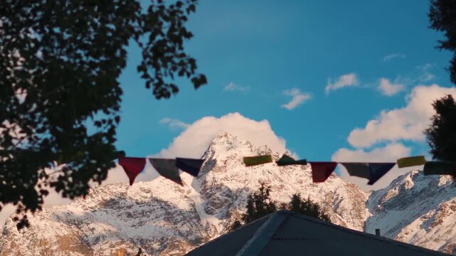 Rack focus shot of prayer flags waving in front of snow covered Himalayan mountain peak during the sunset as seen from Keylong in Lahaul and Spiti district, Himachal Pradesh, India.