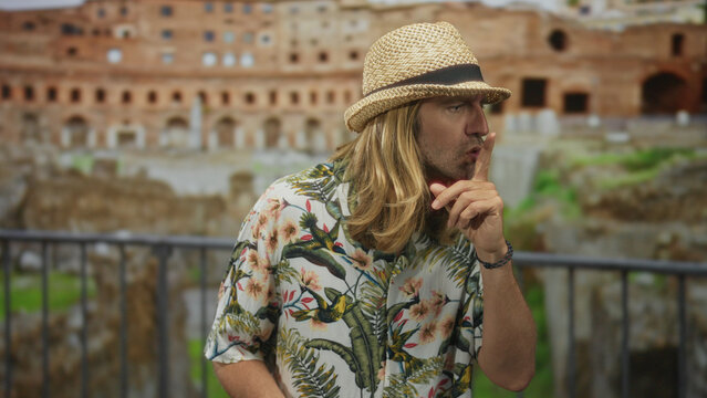 Man with long blonde hair and straw hat holds finger to lips amid ruins inside an ancient colosseum building; silence.