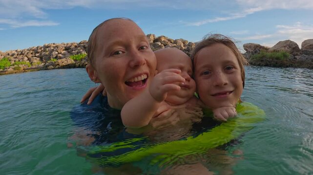Happy mother hugging her two sons standing in the sea, joyful family moment during summer vacation. Tender bonding with ten year old boy and infant. Slowmotion video.