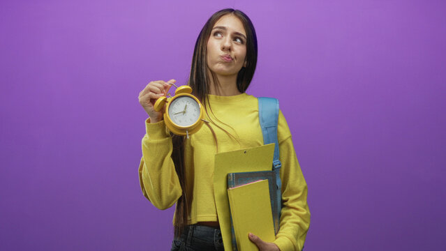 Teenage student holds yellow alarm clock and notebooks, pouts while carrying backpack in studio; time pressure frustration.