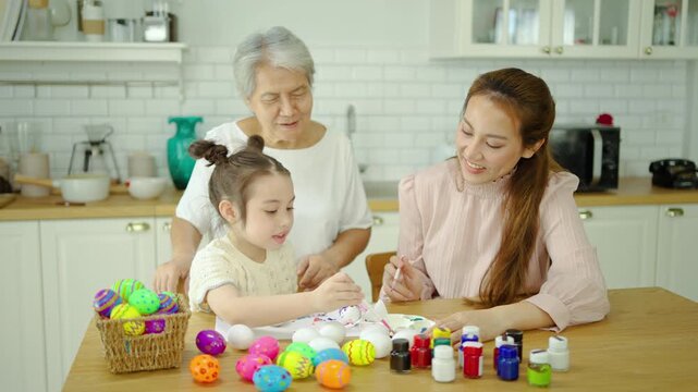 Happy multi-generation Asian family painting Easter eggs together in a bright modern kitchen. Grandmother, mother, and cute little daughter enjoy creative holiday craft activities and bonding.