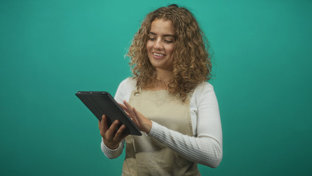 Woman in apron tapping tablet with right hand and holding device with left in studio, smiling while gesturing to screen; digital learning joy.