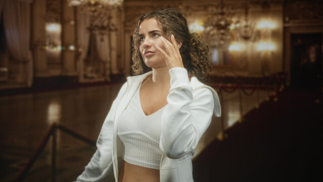 Woman touches forehead while brushing curly hair, navel visible in cropped top inside ornate building with chandeliers and ropes; quiet contemplation.