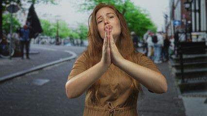 Fototapeta premium Woman in brown dress with hands pressed in prayer gesture on city street amid blurred pedestrians; prayer hope.