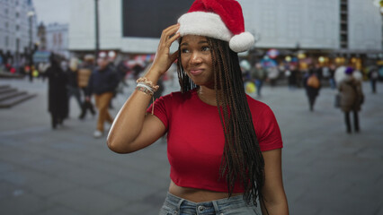 Fototapeta premium Woman touching santa hat on busy street, hand to head, red crop shirt with exposed midriff and bracelets; holiday playful.