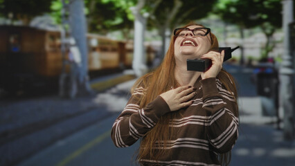 Fototapeta premium Redhead woman laughing while holding a large cellphone on street with hand on chest and wearing glasses; joyful surprise.