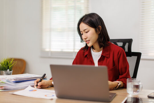 Young Asian woman working from home, focused on laptop while analyzing financial spreadsheets and paperwork at a wooden desk, taking notes and reviewing documents