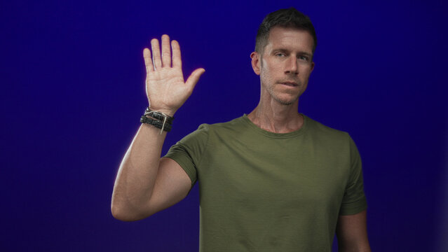 Man raising left hand with palm visible and wristwatch, wearing olive tshirt in deep blue studio; greeting acknowledgement calm.