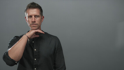 Obraz premium Man with hand to neck gesture showing bare forearm and direct gaze in studio with a grey wall; pensive reflection resolve.