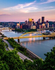 Obraz premium Skyline view of a city at sunset with multiple bridges over a river, showcasing a vibrant sky. Green foliage frames the image