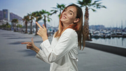 Fototapeta premium Young redhead woman in white coat pointing finger guns and smiling on a street waterfront with palm trees and boats; playful.