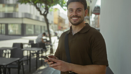 Fototapeta premium Man in brown polo shirt holding smartphone and typing with thumbs while standing on a street terrace outdoors, smiling at the screen; happiness connection.