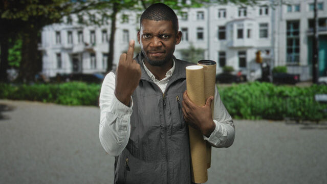 Man holding rolled blueprints shows middle finger toward camera in street with stern expression and vested shirt, standing near city buildings; defiance.