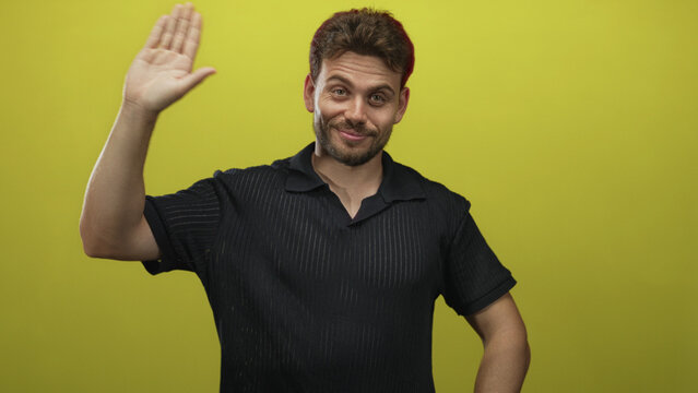 Man raising right palm clearly visible against bright yellow studio wall, wearing black polo shirt with casual stance and slight smile; friendly greeting.