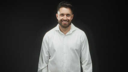 Obraz premium Man with beard smiling, hands at sides in studio wearing white shirt and collar visible; friendly approachable presence.