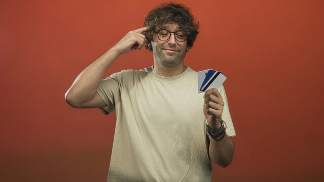 Man holding creditcard, points finger to temple in studio with orange backdrop; thoughtful decision pose.