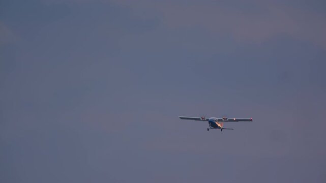 A twin-engine propeller airplane with navigation lights on flies and banks in a graceful turn against a muted, overcast sky during twilight or a cloudy day