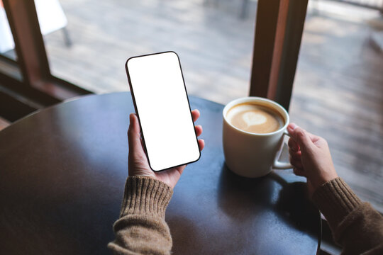 Mockup image of a woman holding mobile phone with blank desktop screen while drinking coffee in cafe