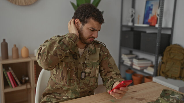 Hispanic man in camouflage uniform holds smartphone and taps screen in building, cringing at message received; stress frustration isolation anxiety.