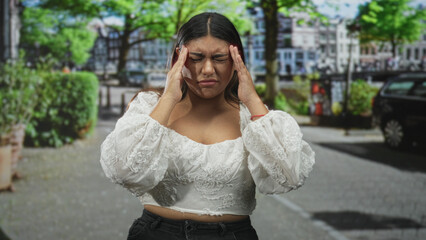 Fototapeta premium Woman pinches nose on street wearing white embroidered cropped top with hand on hip and eyes closed, near parked car and hedge; discomfort concern.