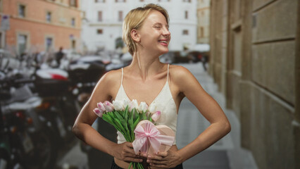 Fototapeta premium Woman holding tulips and a heart giftbox, presenting the flowers with smiling hands on a street lined with scooters and buildings; romantic joy.