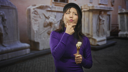 Obraz premium Woman holding makeup brushes, finger to chin gesture, posed before stone sculptures in building; quiet contemplation.