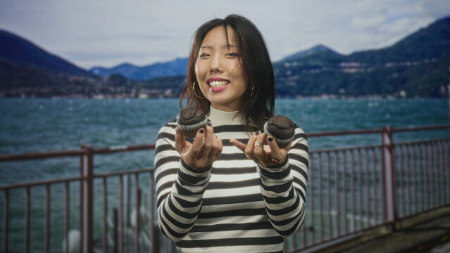 Woman holding chocolate muffin, offering with one hand by waterfront railing on street; playful curiosity.