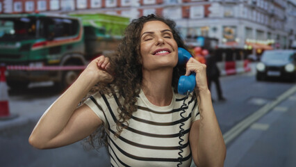 Fototapeta premium Woman holding bright blue telephone receiver brushing hair in front of construction barriers on a city street; joy.