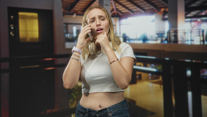 Fototapeta premium Woman wearing headphones holds phone to ear with hand on chin and exposed stomach in an airport terminal; anxiety concern.