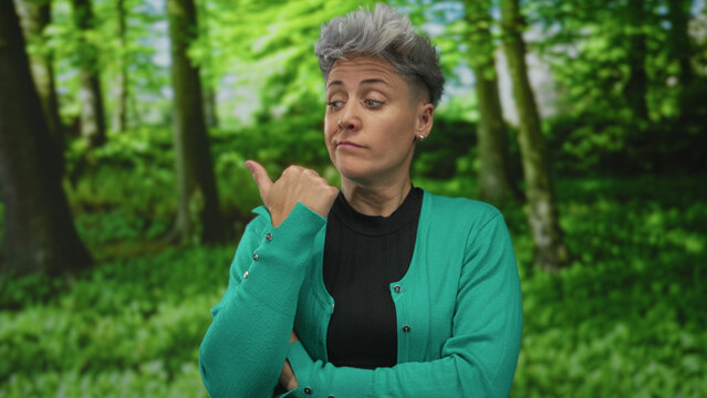 Woman with short gray hair wearing green cardigan and black top points thumb at forest backdrop indoors; skepticism doubt cynicism distrust.