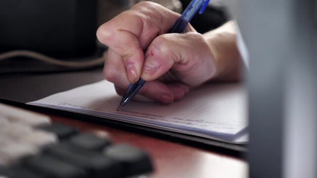 A close-up of the hand of a woman with nail dystrophy writing with a pen in a notebook while sitting at a table.
