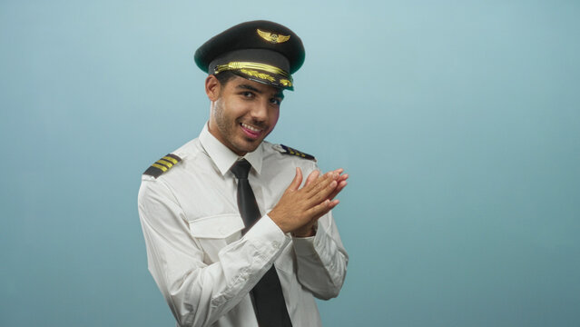 Man pilot in captain cap and white uniform clasps hands and smiles, epaulettes visible against a pale blue studio backdrop; playful confidence.