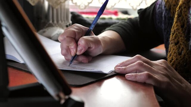 A close-up of the hand of a woman with nail dystrophy writing with a pen in a notebook while sitting at a table.