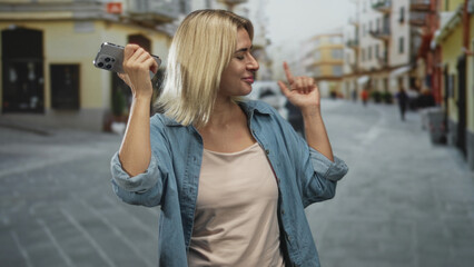 Fototapeta premium Young woman holding smartphone to ear and smiling with a playful raised hand on a cobblestone street; contentment wanderlust.