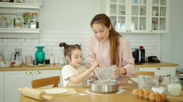 Happy Asian mother and cute little daughter baking together in a bright modern home kitchen. Mom helps the girl sift flour into a bowl to make dough. Beautiful family bonding and fun cooking times.