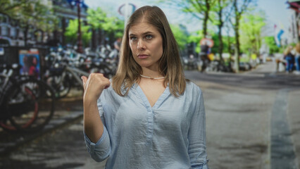 Fototapeta premium Blonde woman pointing thumb to street beside sunlit canal flanked by bicycles in amsterdam; skepticism.