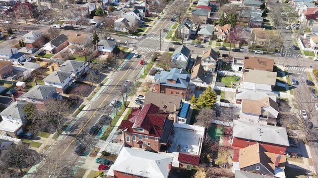 An angled aerial perspective shows closely arranged houses, rooftops and streets bordered by green trees in a residential neighborhood. The roofs form a distinct pattern within the leafy neighborhood.