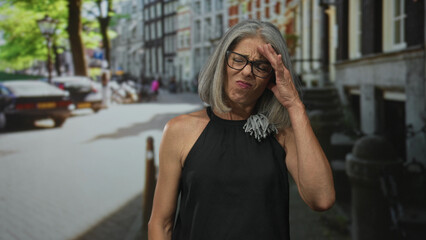 Fototapeta premium Woman with hand on forehead on street in city wearing glasses and black sleeveless dress; concern uncertainty.