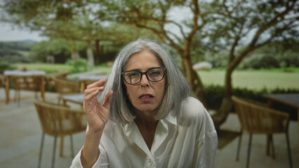 Fototapeta premium Woman grey haired adjusting eyeglasses while squinting and leaning forward on a restaurant terrace outdoors; curiosity reflection.