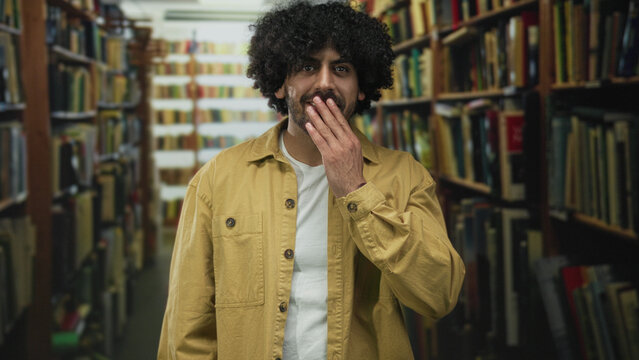 Man with hand to mouth standing in a library building aisle among bookshelves, wearing jacket and tshirt, mouth open; surprise curiosity.