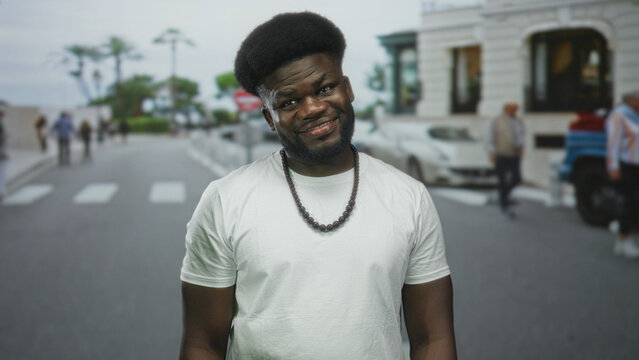 Man smiling with visible face and beaded necklace on a city street crosswalk with blurred pedestrians and cars; confidence charisma ease.