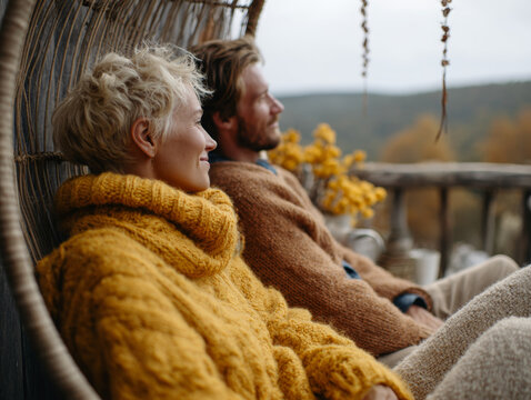 A man and a woman in cozy sweaters spend quality time at their country home, gazing at the bright autumn landscape filled with golden and red foliage. Serene lifestyle.