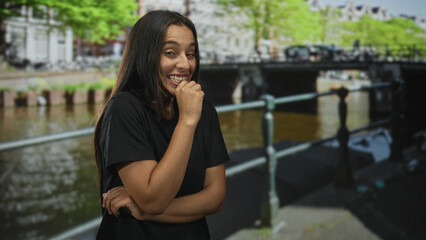 Fototapeta premium Woman laughing with fist to mouth and crossed arms standing by canal railing on an amsterdam street with bridge behind her; joy spontaneity.
