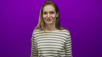 Fototapeta premium Young caucasian woman waves hand in purple studio setting with bright lighting and casual striped shirt visible; welcome happiness friendliness warmth.