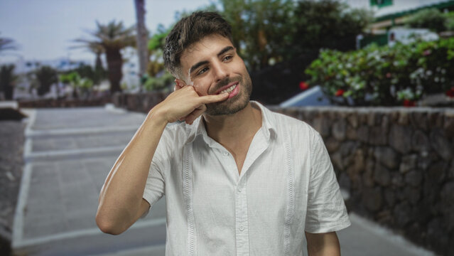 Man wearing white linen shirt making call gesture with hand on city street; friendly invitation to connect.