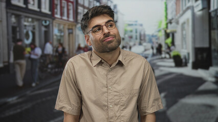 Obraz premium Man wearing glasses tilts head slightly with closed eyes in a busy city street lined with brick storefronts and blurred pedestrians; indifference.