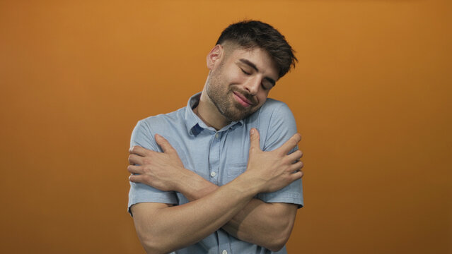 Man crosses arms over chest hugging himself with closed eyes and a smile in orange studio; serenity.